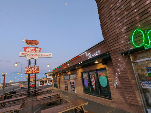 Exterior of Del's Restaurant at dusk, featuring a cow sign, neon lights, and outdoor seating.